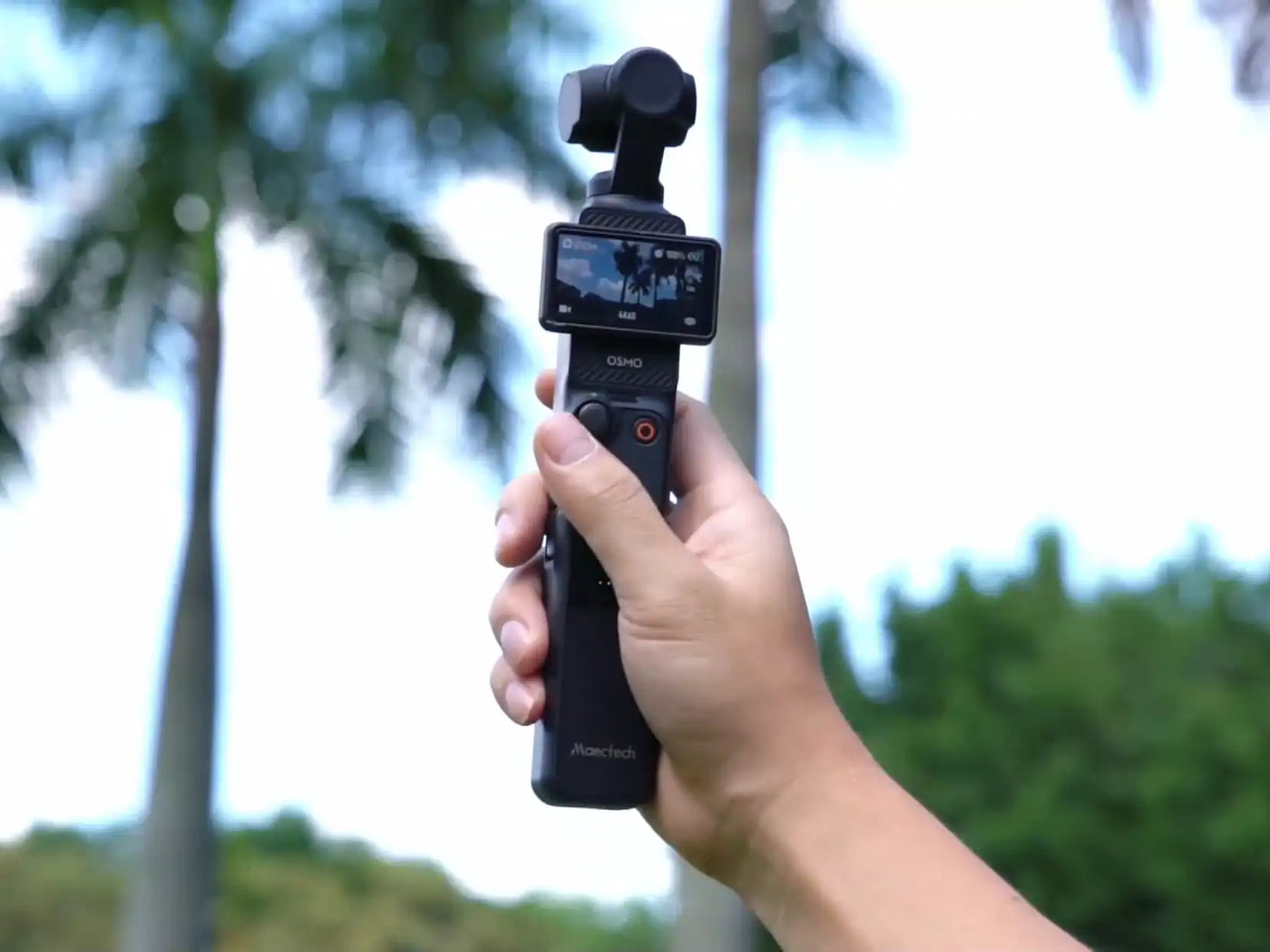 Hand holding a small camera with a screen, blurred green outdoor background